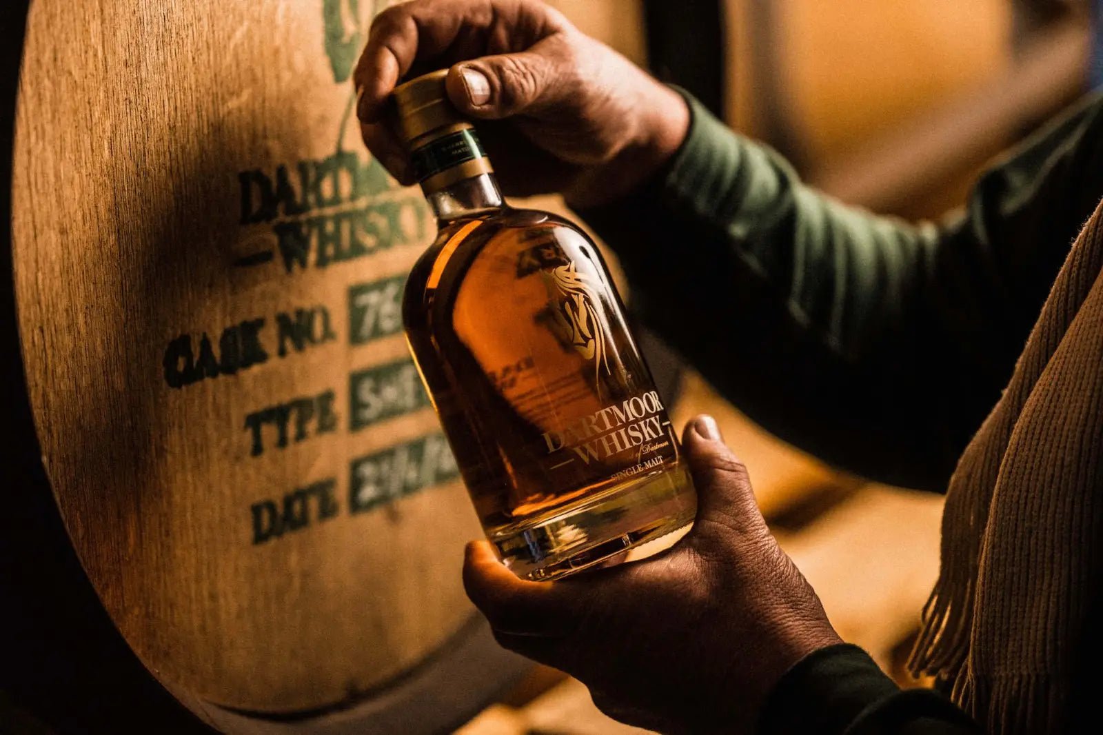 Hands holding a bottle of Dartmoor whisky in front of a barrel