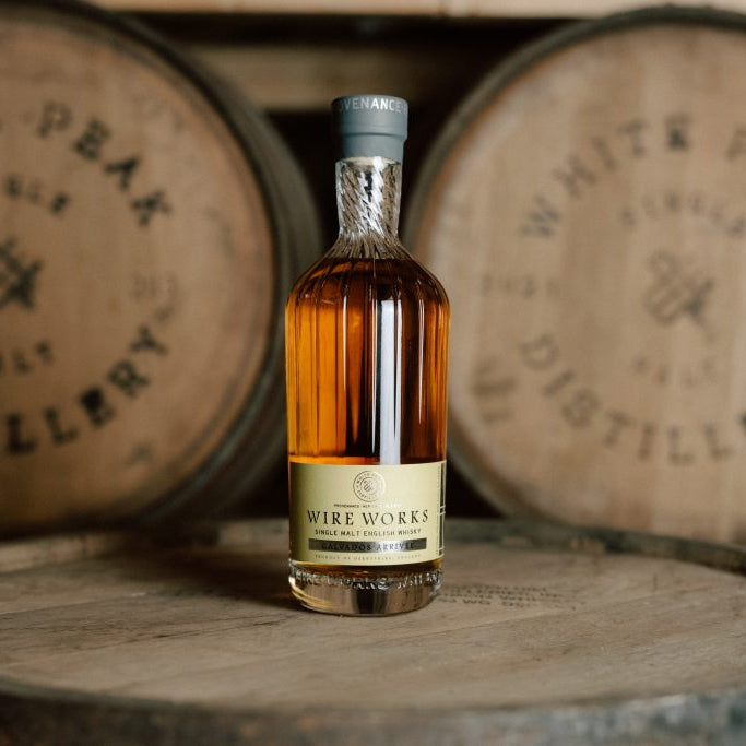 A bottle of Wire Works Calvados Arrivee english whisky stands on a wooden barrel with two blurred White Peak Distillery barrels in the background.