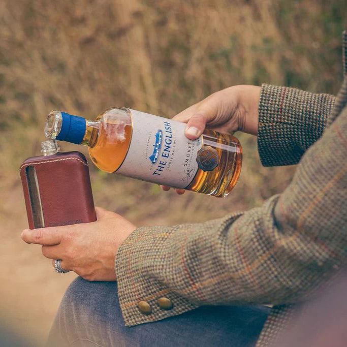 Person in tweed jacket holding a bottle of The English Whisky Company Smokey whisky and a leather flask outdoors.