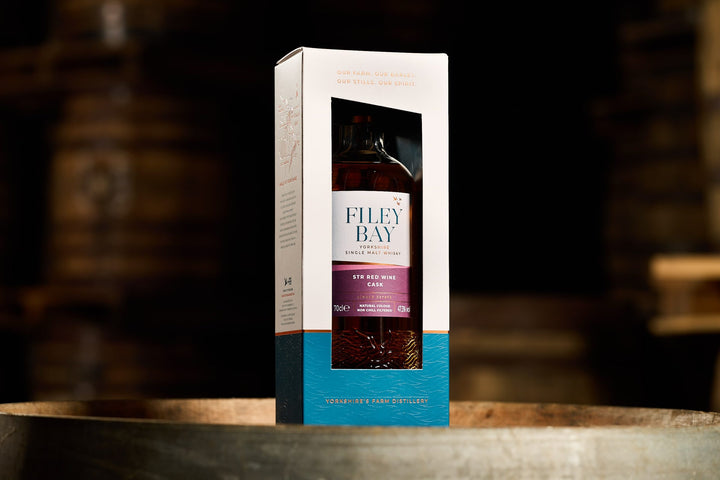 Filey Bay Flagship STR Red Wine Single Malt English whisky bottle and box on a wooden surface with a blurred background