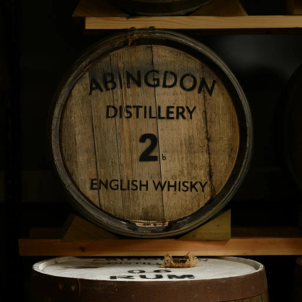 A wooden whisky barrel with 'ABINGDON DISTILLERY 2 B ENGLISH WHISKY' printed on the barrel resting on a wooden shelf in a dark storage room.