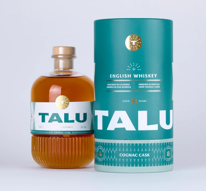 A bottle of TALU English Whiskey with amber liquid and a white label, standing next to its teal cylindrical packaging tube on a light gray background.