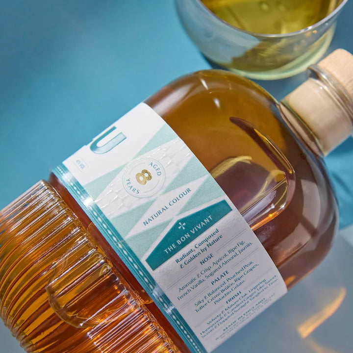 A bottle of Taste Talu Cognac Cask Whiskey filled with golden-amber liquid and featuring a white and teal label with a wooden cap, set against a blue background with a glass of whisky in the background.