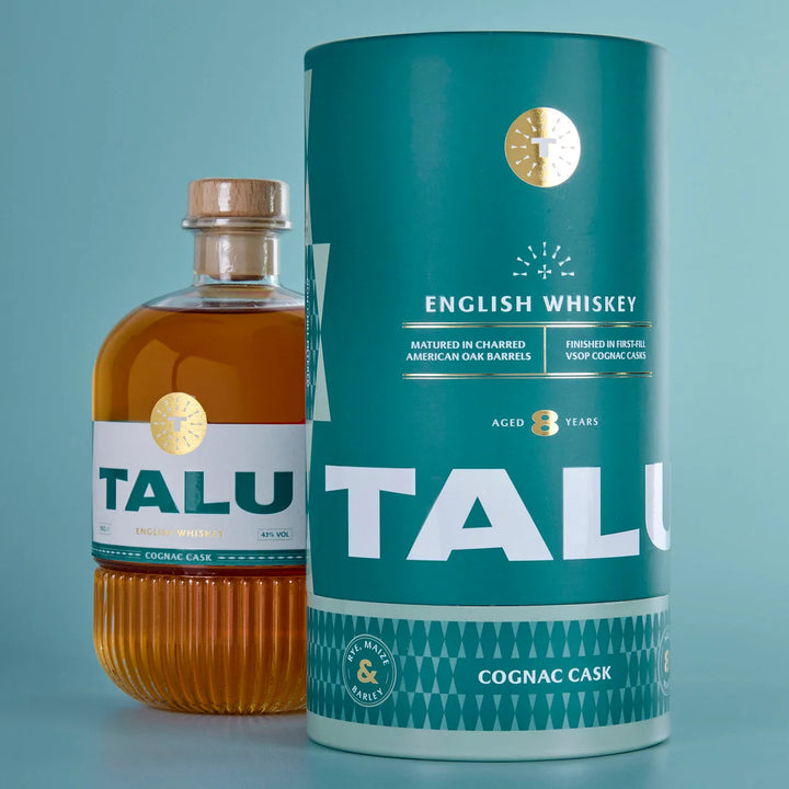 A bottle of Talu English Whiskey next to its cylindrical teal packaging container, both featuring a gold seal and white branding, set against a solid light blue background.