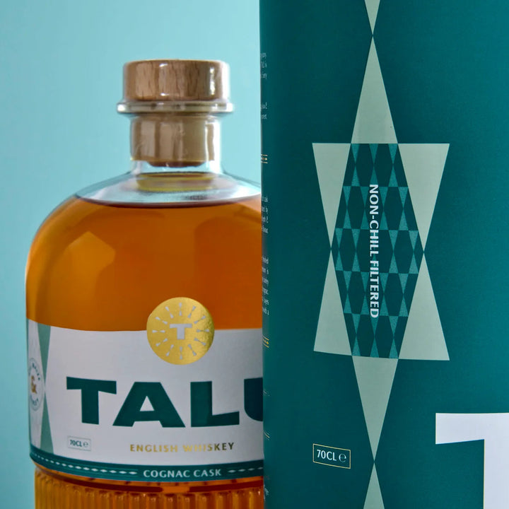 A bottle of TALU English Whiskey Cognac Cask whiskey with amber liquid, a wooden stopper, and a white label, shown next to its teal packaging box with geometric patterns.