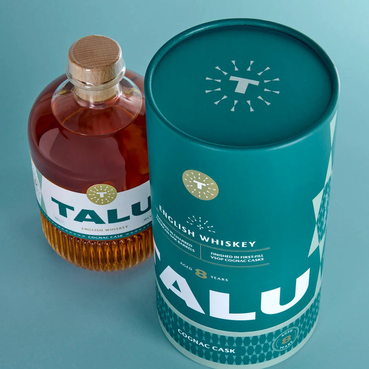 A bottle of Talu English Whiskey next to its teal cylindrical tin packaging on a light blue background.