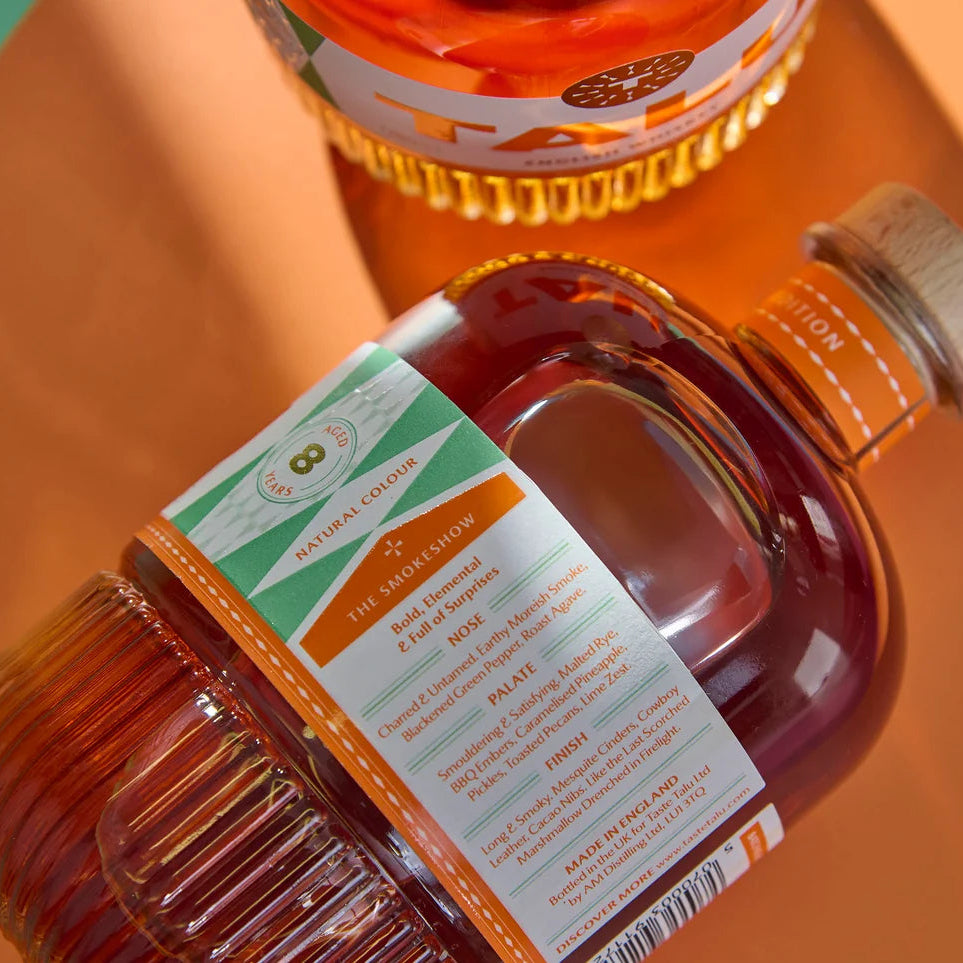 A close-up of a bottle of Talu Mezcal Cask English Whiskey with a white, orange, and green label, lying diagonally on a warm orange surface. Another bottle is partially visible in the background.
