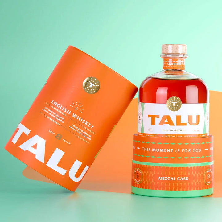 A bottle of Talu English Whiskey with a clear glass body showing amber liquid, next to its matching orange cylindrical packaging sleeve, set against a mint green and orange background.