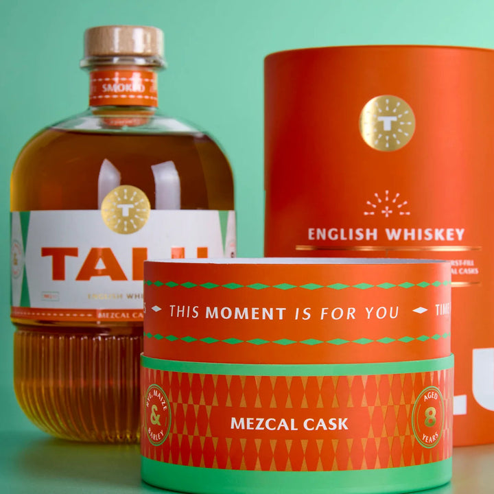A bottle of Talu Mezcal Cask whiskey alongside its custom orange and green packaging, including a cylindrical box and a tin canister, set against a solid mint green background.
