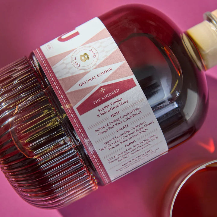 A bottle of Talu Oloroso Cask English Whiskey with a ribbed glass body and a cork stopper, shown diagonally on a pink background with a glass of the red liquid partially visible in the lower right.