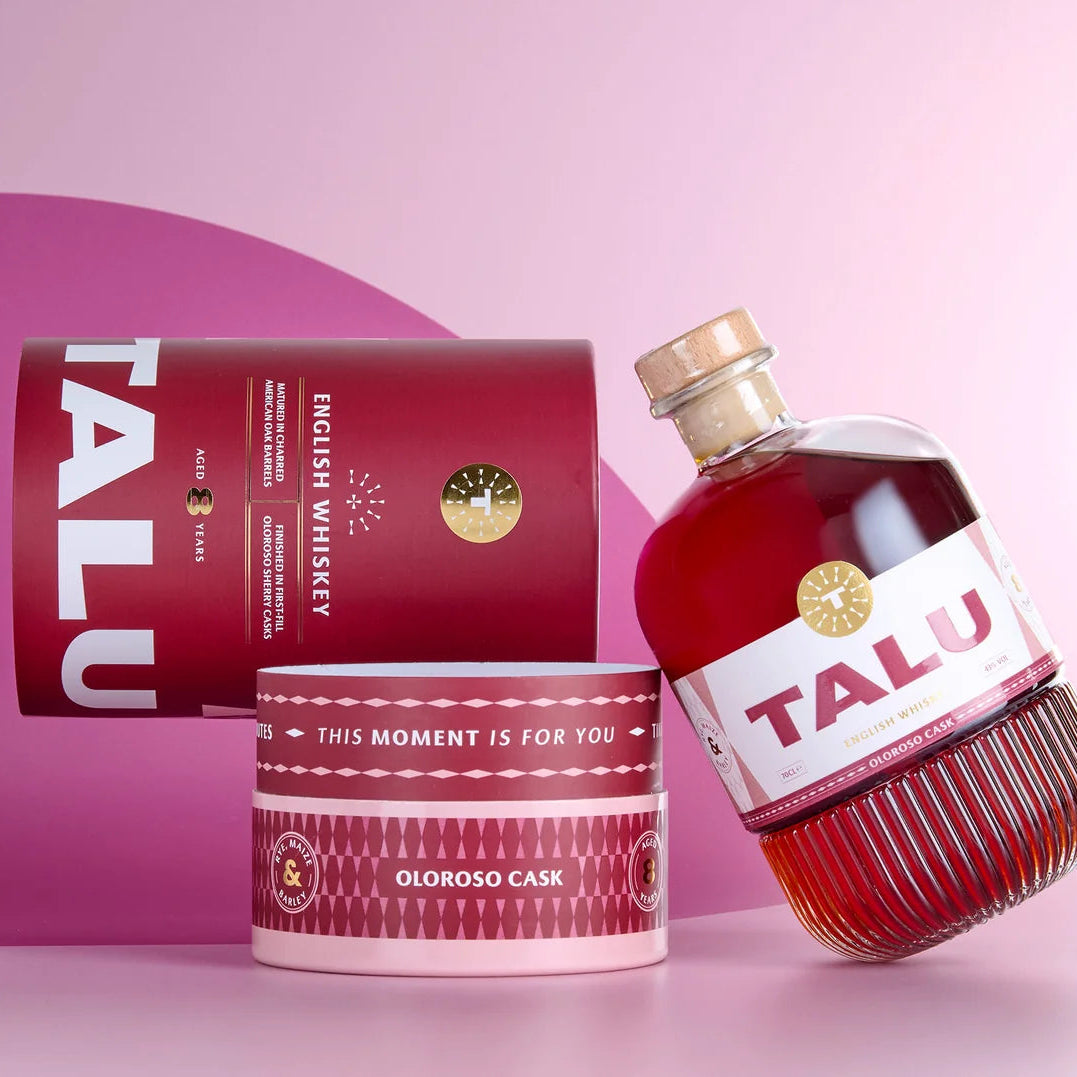 TALU Oloroso Cask English Whiskey bottle and packaging set on a pink studio background. The clear glass bottle contains deep red whiskey and has a white label with red TALU lettering.