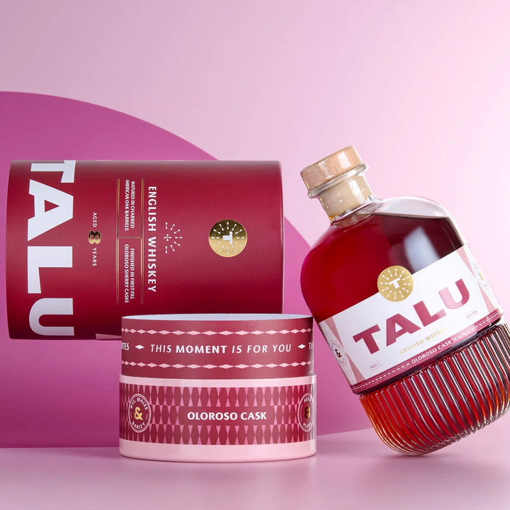 TALU Oloroso Cask English Whiskey bottle and packaging set on a pink studio background. The clear glass bottle contains deep red whiskey and has a white label with red TALU lettering.