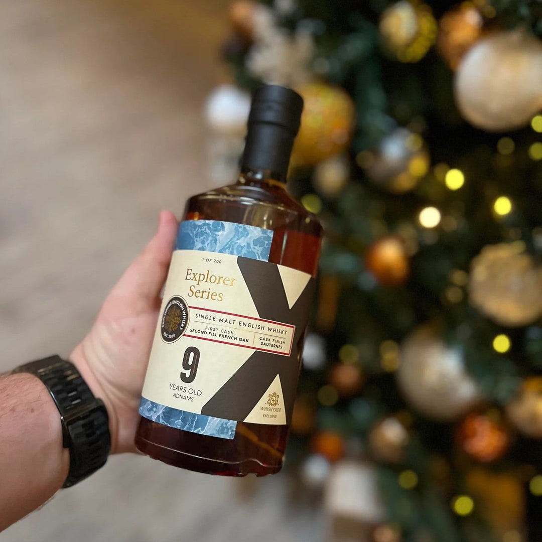 Hand holding a bottle of 'Explorer Series 9 Year Old' whisky in front of a decorated Christmas tree.