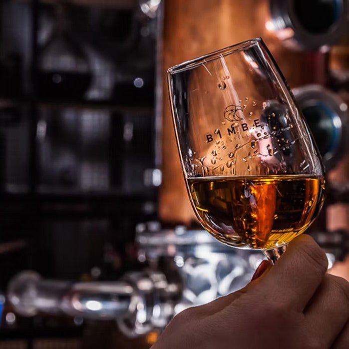 A hand holds a clear glass of whisky in front of a blurred distillery background with copper vessels.