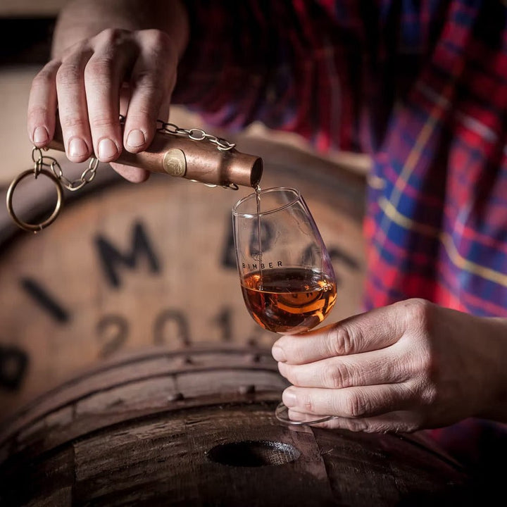 A person in a red and blue checked shirt pours whisky into a small glass, with a wooden barrel blurred in the background.
