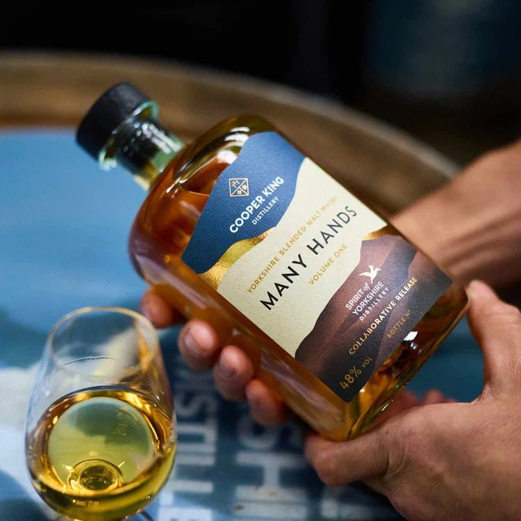 A hand holds a bottle of Cooper King Many Hands Yorkshire whisky next to a small glass of whisky, with a blurred barrel top background