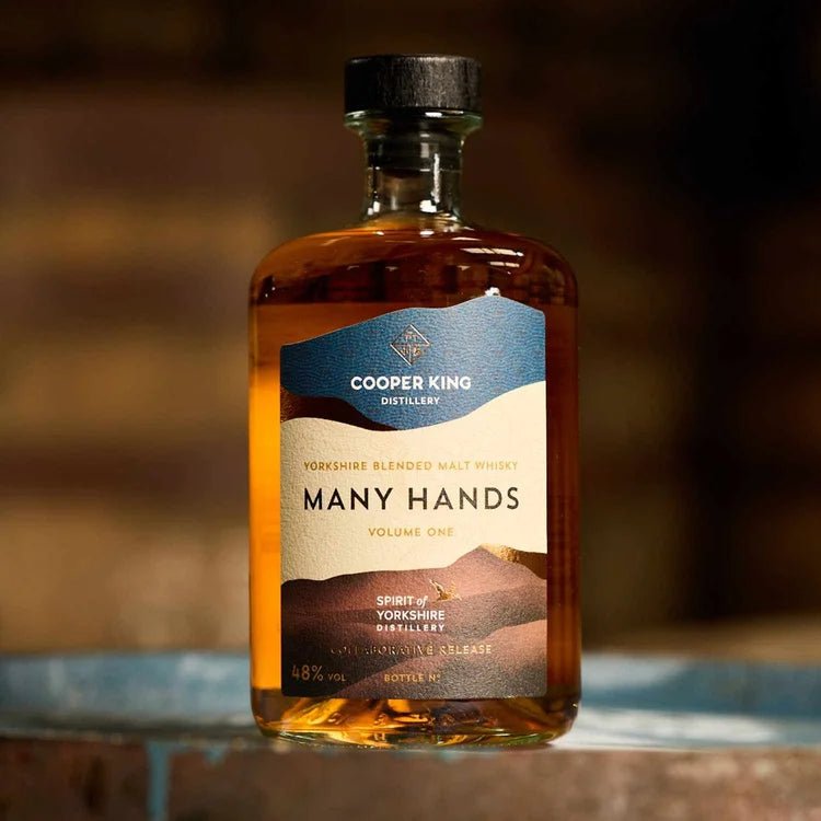 A bottle of Cooper King Distillery Many Hands Yorkshire Blended Malt whisky, showing the whisky inside a glass bottle with a blue and beige label, set against a warm, blurred background