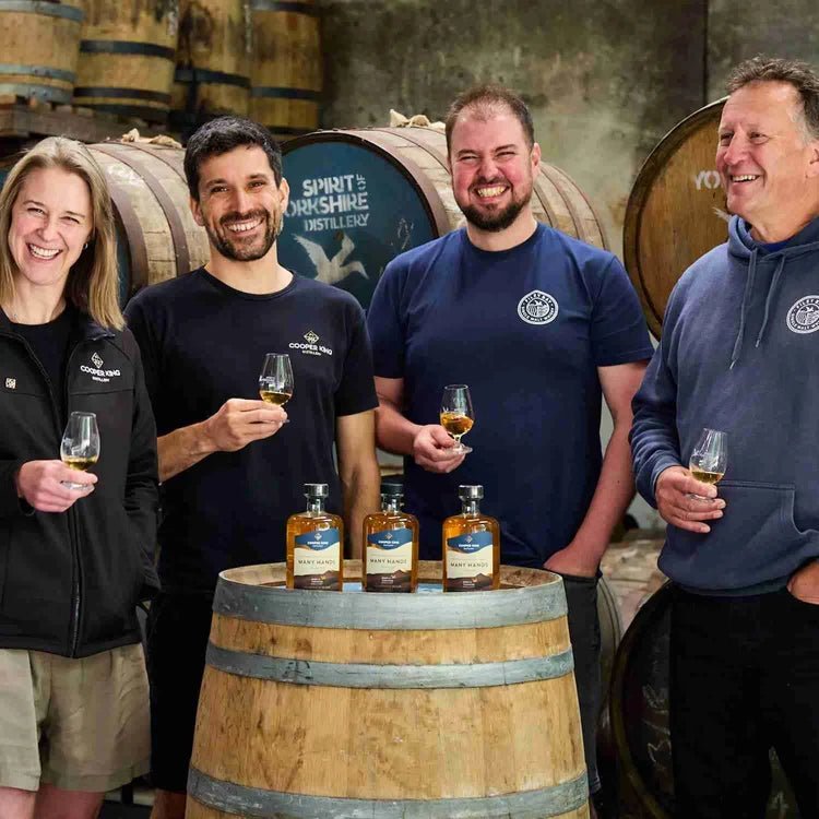 Four people standing in a whisky distillery holding whisky glasses, with whisky bottles displayed on a wooden barrel in the foreground