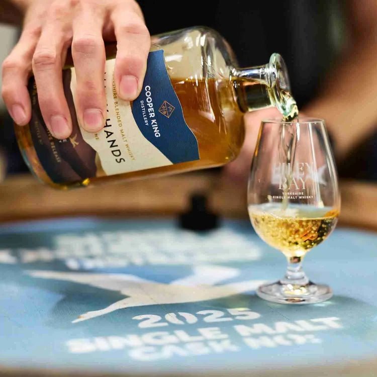 A hand pours amber whisky from a bottle labeled Cooper King Many Hands Malt into a glass, set on a blue painted barrel top
