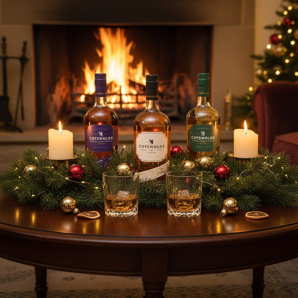 Three bottles of Cotswolds whisky on a table with glasses and Christmas decorations in front of a fireplace.