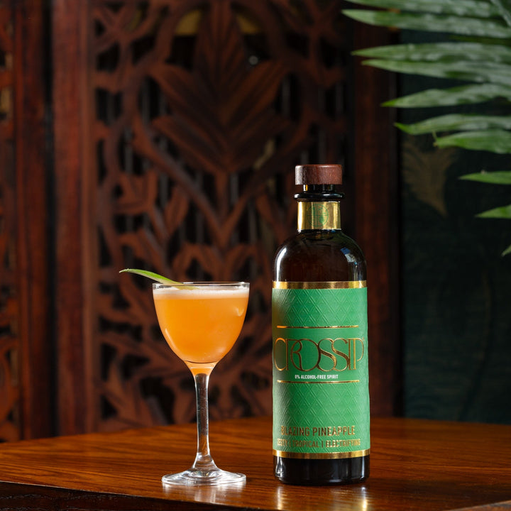 Bottle of Crossip Blazing Pineapple on a wooden table with a cocktail glass filled with an orange drink.