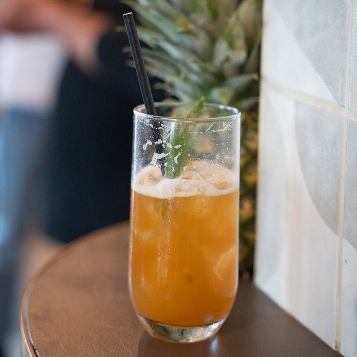 Glass of Crossip Blazing Pineapple cocktail with ice and a straw on a wooden surface, with a blurred background.