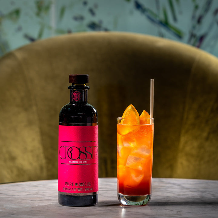Bottle of Crossip Pure Hibiscus with a cocktail garnished with orange slices on a table.