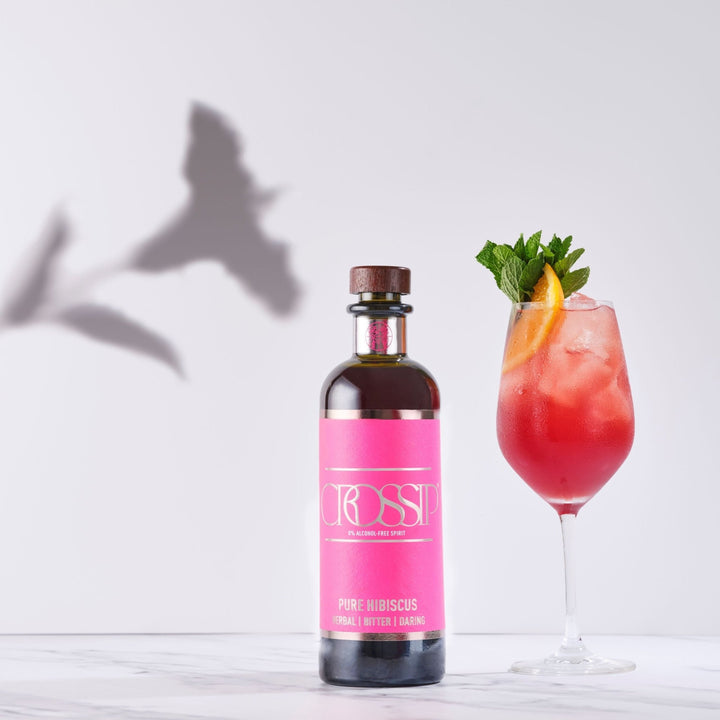 Bottle of Crossip Pure Hibiscus with a pink cocktail garnished with mint and lemon on a white background