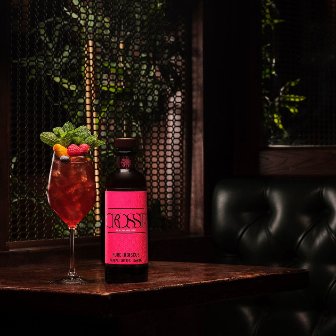 Cocktail with fruit garnish and a bottle of Crossip Pure Hibiscus on a wooden table with a dark background.