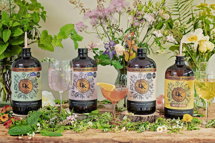 Four bottles of Drinks Kitchen non-alcoholic botanical aperitifs with decorative flowers and glasses on a wooden table.