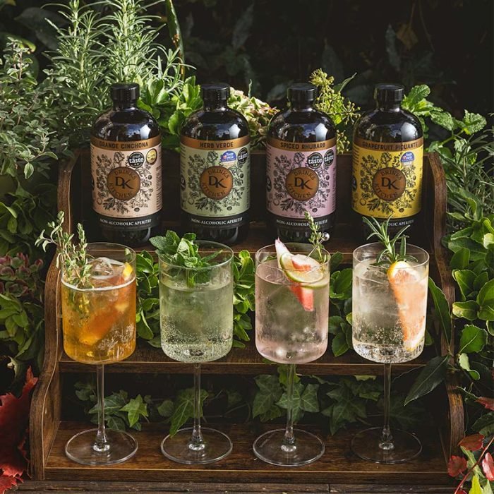 Four bottles of Drinks Kitchen non-alcoholic botanical aperitifs with four glasses garnished with herbs and fruits on a wooden stand with greenery in the background.