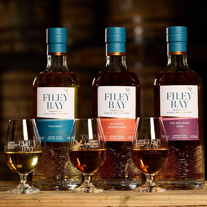 Three bottles of Filey Bay whisky with corresponding glasses on a wooden surface.