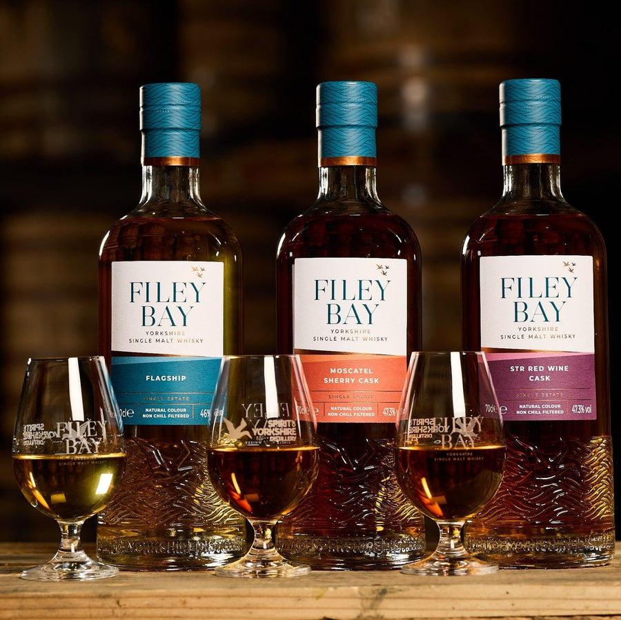 Three bottles of Filey Bay whisky with corresponding glasses on a wooden surface.
