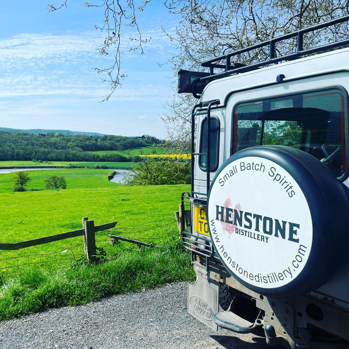 Landrover with Henstone Distillery logo parked by a scenic landscape with a river and green fields.