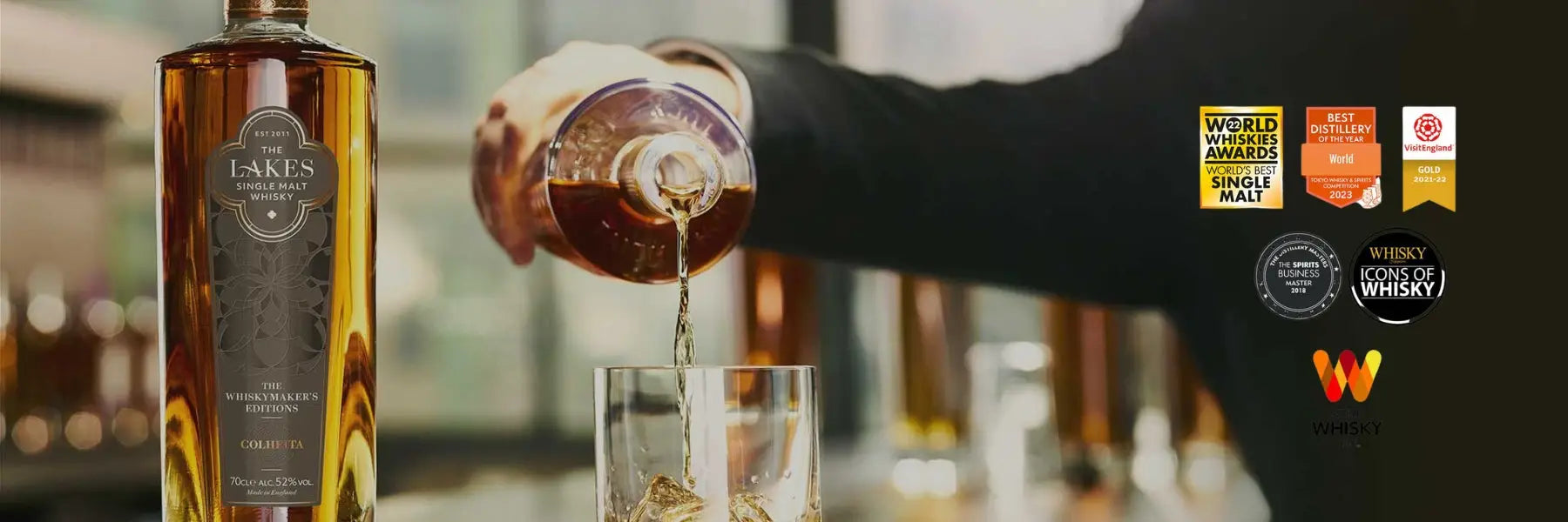 Person pouring whiskey into a glass with a bottle of Lakes whisky in the foreground, surrounded by awards.
