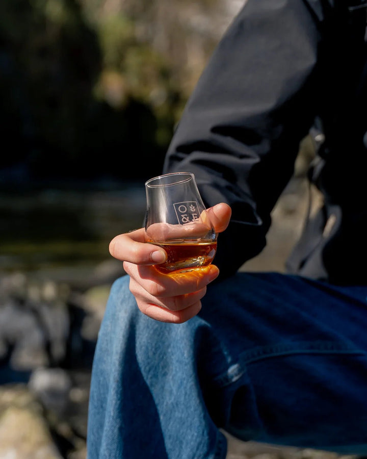 Person holding an Oak &amp; Ember nosing glass of whisky liquid outdoors with a blurred natural background