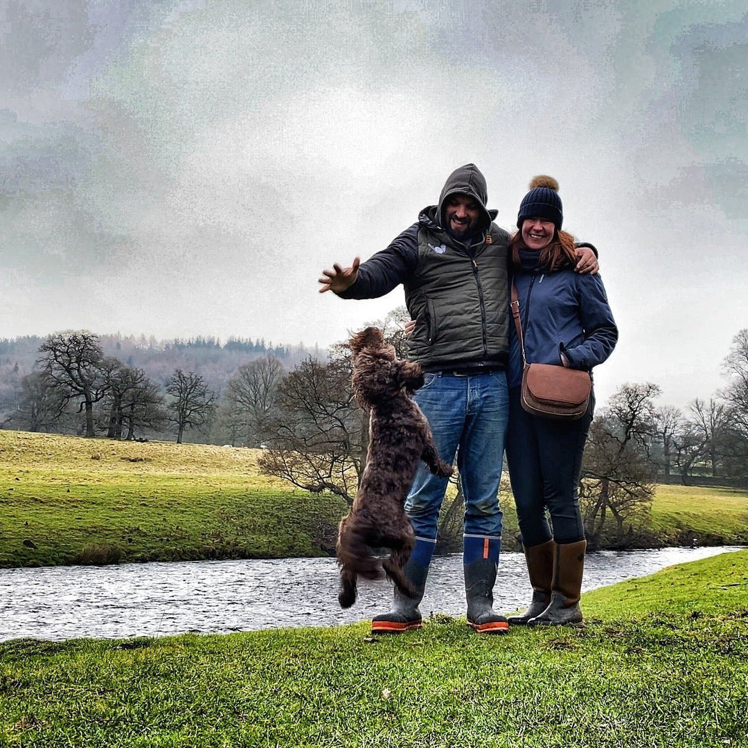 Two people and a dog standing on a grassy area near a river with trees in the background.