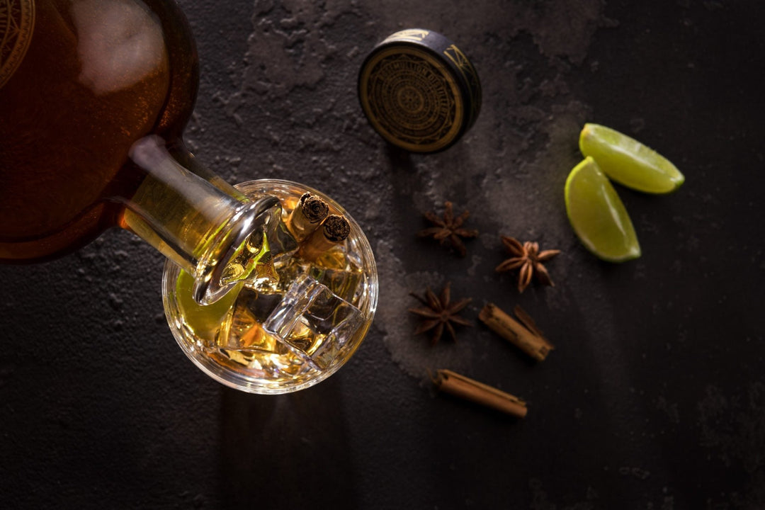 Bottle of rum with a glass filled with rum and ice, surrounded by spices and lime wedges on a dark surface.