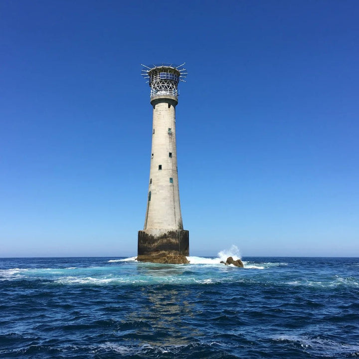 Scilly Isles Lighthouse on a rocky outcrop in the middle of the ocean with clear blue sky.