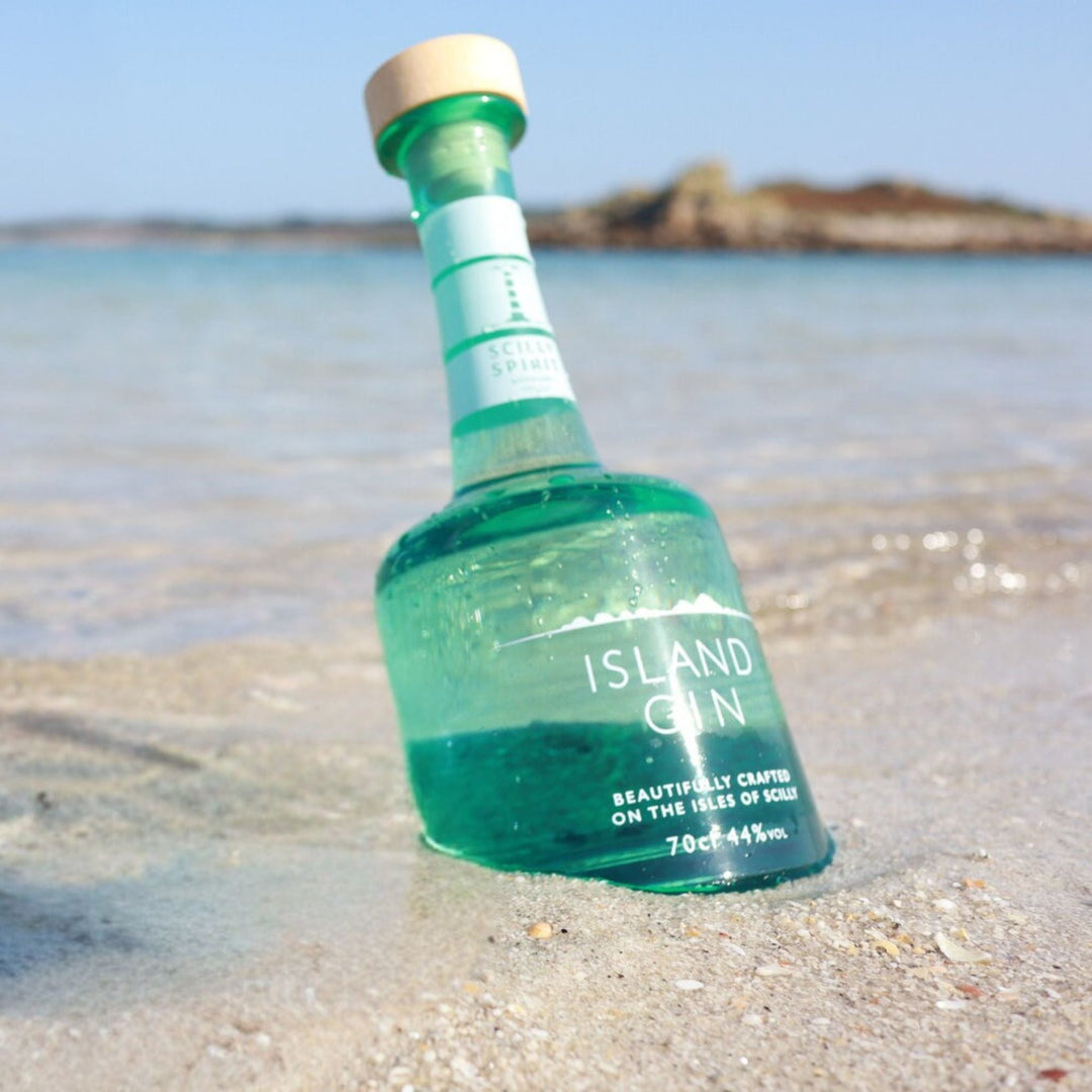 Green bottle of Scilly Spirit Island Gin on a sandy beach with water and sky in the background