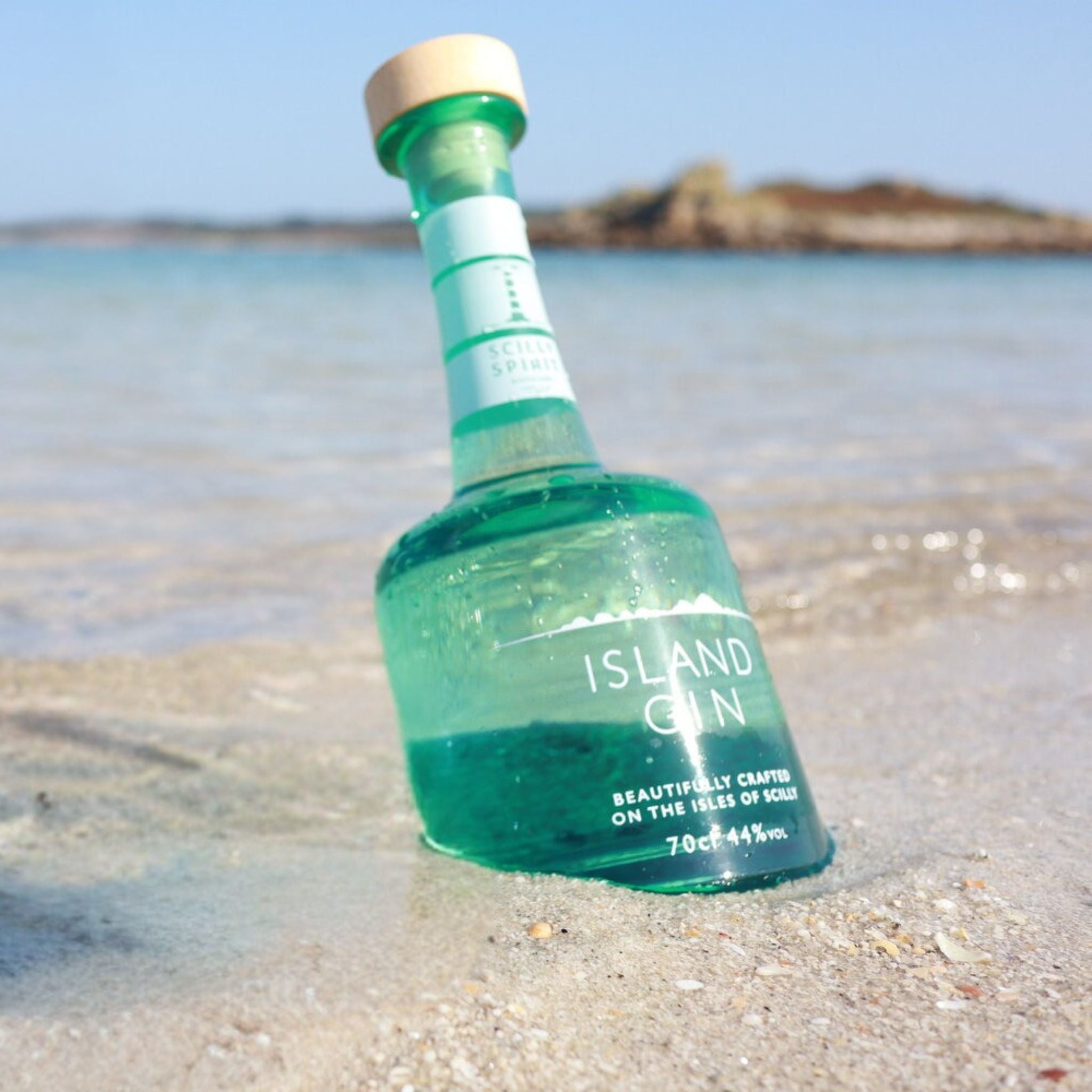 Green bottle of Scilly Spirit Island Gin on a sandy beach with water and sky in the background