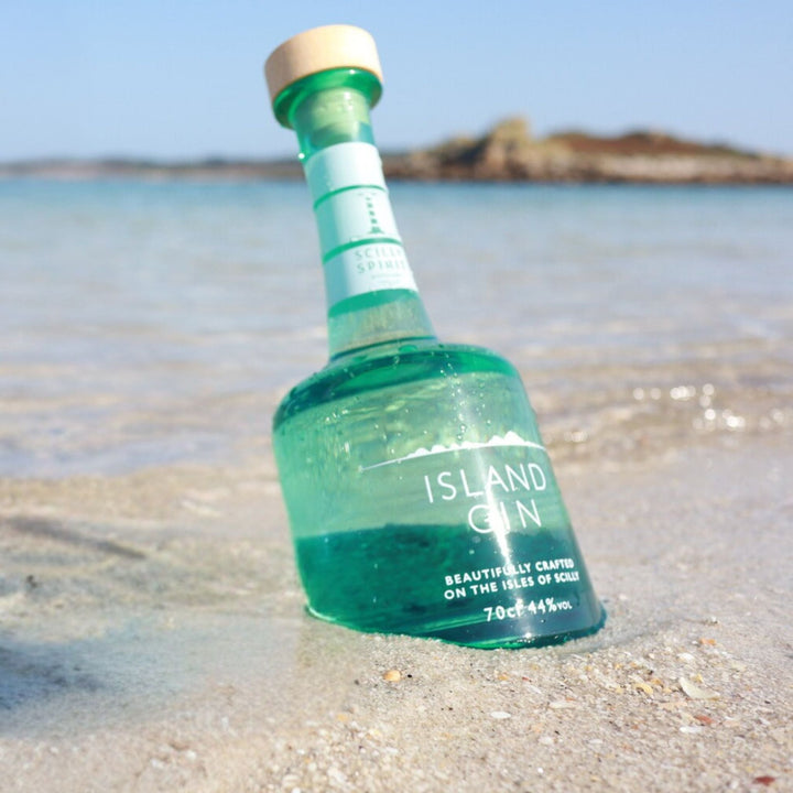 Green bottle of Scilly Spirit Island Gin on a sandy beach with water and sky in the background