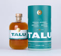A bottle of TALU English Whiskey with amber liquid and a white label, standing next to its teal cylindrical packaging tube on a light gray background.
