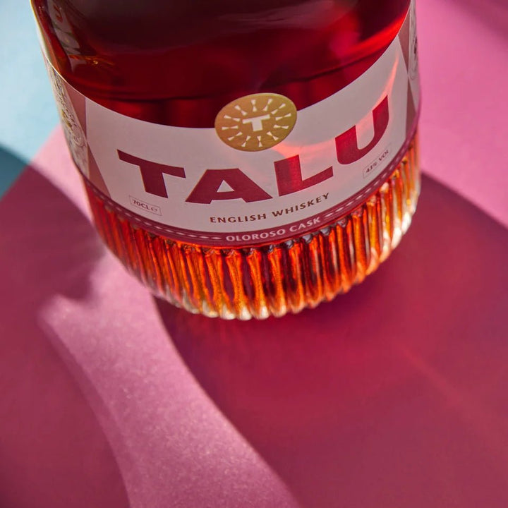 A close-up of a TALU Oloroso Cask English Whiskey bottle showing the amber liquid and part of the white label, set against a vibrant pink background with dramatic shadows.