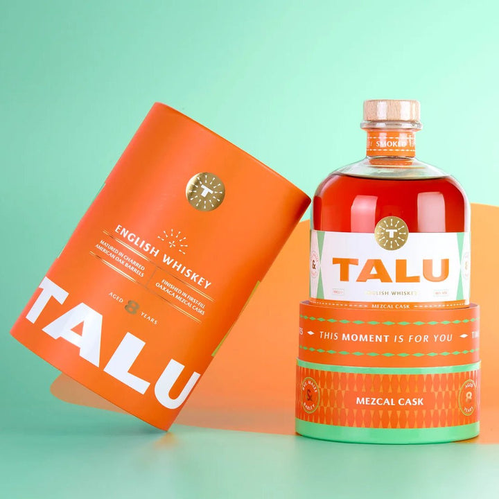 A bottle of Talu English Whiskey with a clear glass body showing amber liquid, next to its matching orange cylindrical packaging sleeve, set against a mint green and orange background.