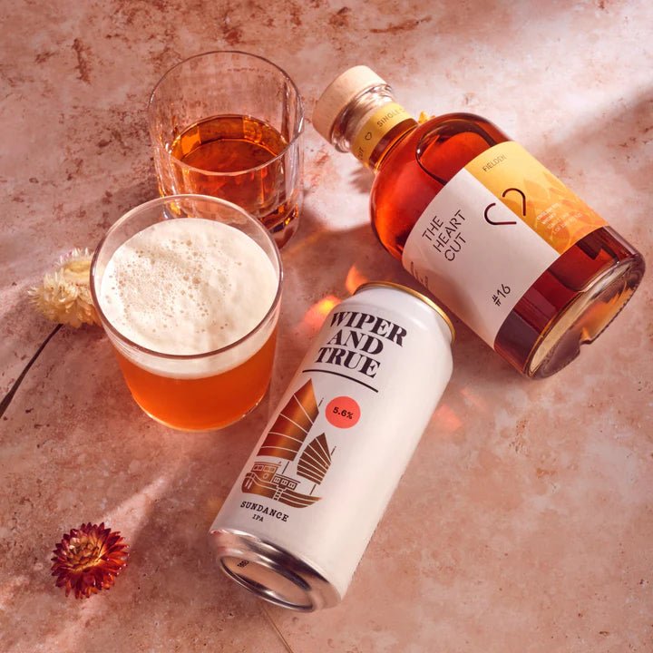 Bottle of The Heart Cut whisky and a can of 'Wiper and True' beer on a textured surface with flowers.