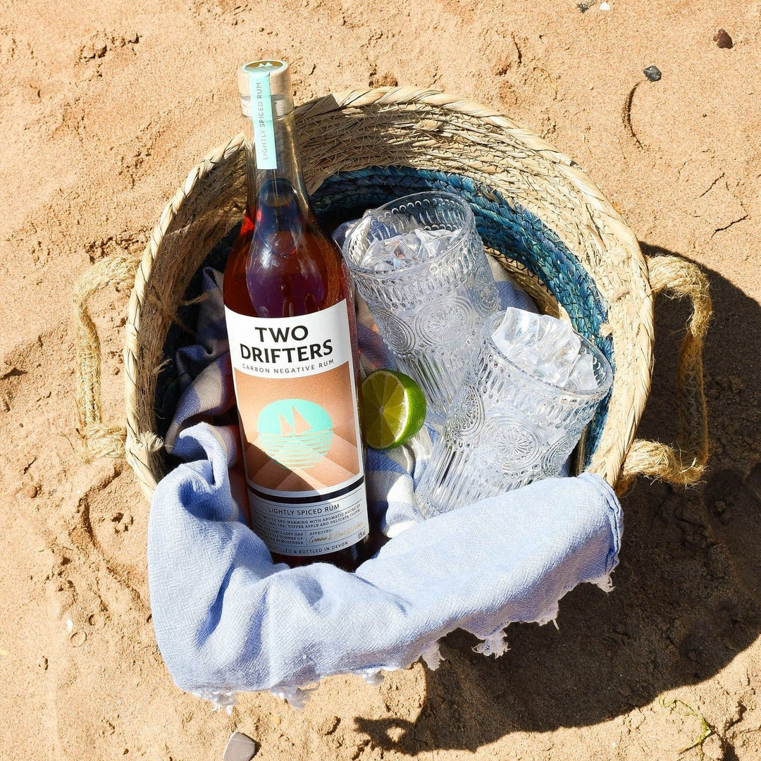 Bottle of 'Two Drifters' lightly spiced rum with ice and a lime in a woven basket on sand