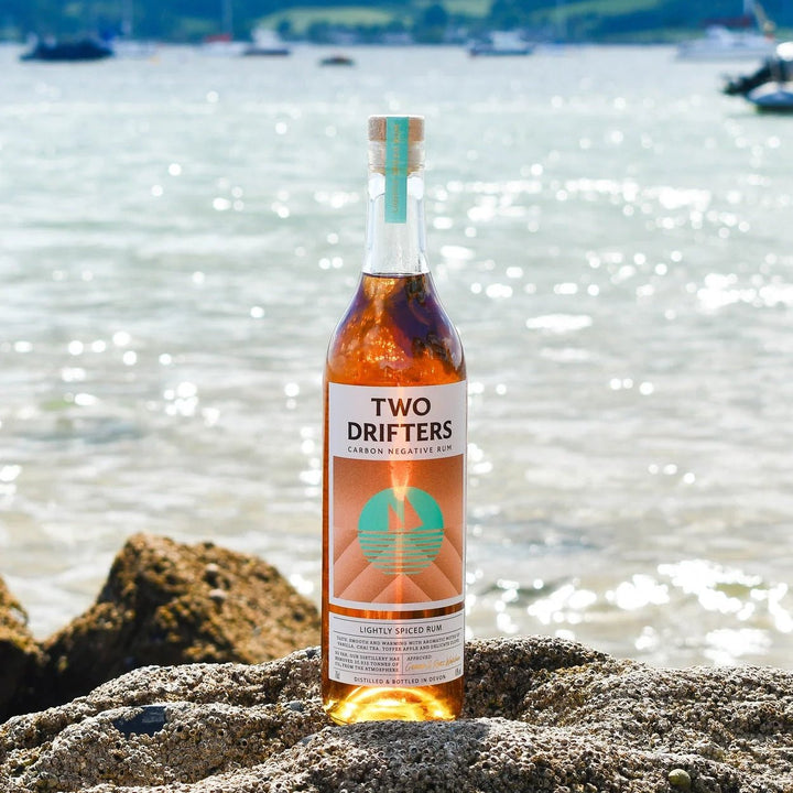 Bottle of 'Two Drifters' rum on a rock by a waterfront with boats in the background