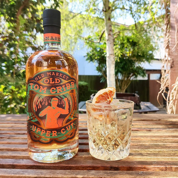 Bottle of Old Market Tom Cribb Upper Cut 50% Gin with a glass of cocktail on a wooden table outdoors.