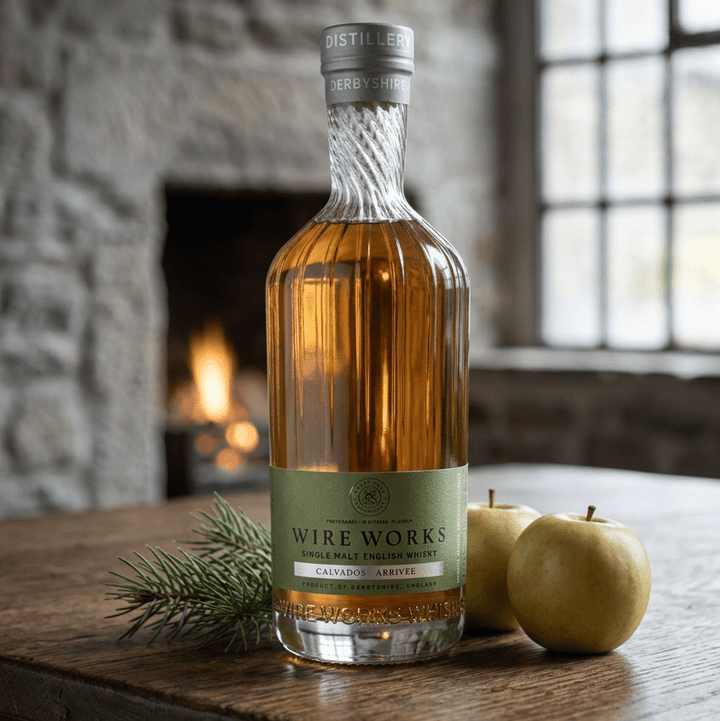 Wire Works Calvados Arrivée single malt whisky on an oak table with green apples and pine with an open fire in the background